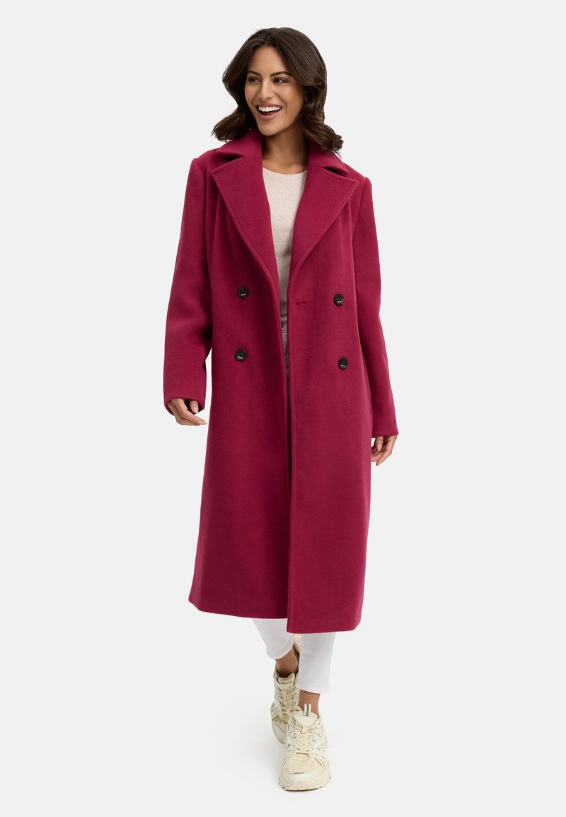 Fuchsia ullkappa med dubbelknäppt design, stor krage och svarta knappar, bärs över en ljus topp och vita byxor, tillsammans med sneakers.