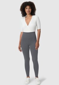 Biały body z krótkim rękawem w formie kopertowej, zestawiony z wysokim stanem i jednolitymi, szarymi legginsami. Modelka ma na sobie białe trampki; widoczne kręcone włosy.