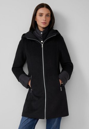 Vrouw draagt een zwarte capuchonjas met twee ritszakken en gelaagde, doorgestikte lange mouwen, staand tegen een grijze achtergrond.