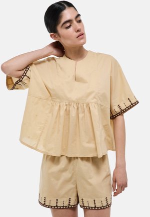 Femme portant un haut beige ample et un short avec une bordure brodée marron, posant avec une main derrière la nuque.