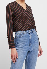 Blusa negra de manga larga con rayas diagonales en color óxido, diseño de cuello en V, combinada con jeans de denim claros de tiro alto.