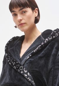 Albornoz negro peludo con ribete de estampado de leopardo y detalles brillantes. Presenta un cuello ancho y un diseño cruzado en la parte delantera. Textura suave y aspecto acogedor.