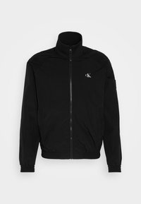 Veste noire à col montant, fermeture éclair pleine longueur et poignets élastiques. Présente un petit logo blanc sur la poitrine gauche. Matière lisse et légère.