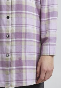 Lila kockás flaneling, fehér és sárga vonalakkal, gombos záródással és puha tapintással. Az ujjak a csuklón túl érnek.