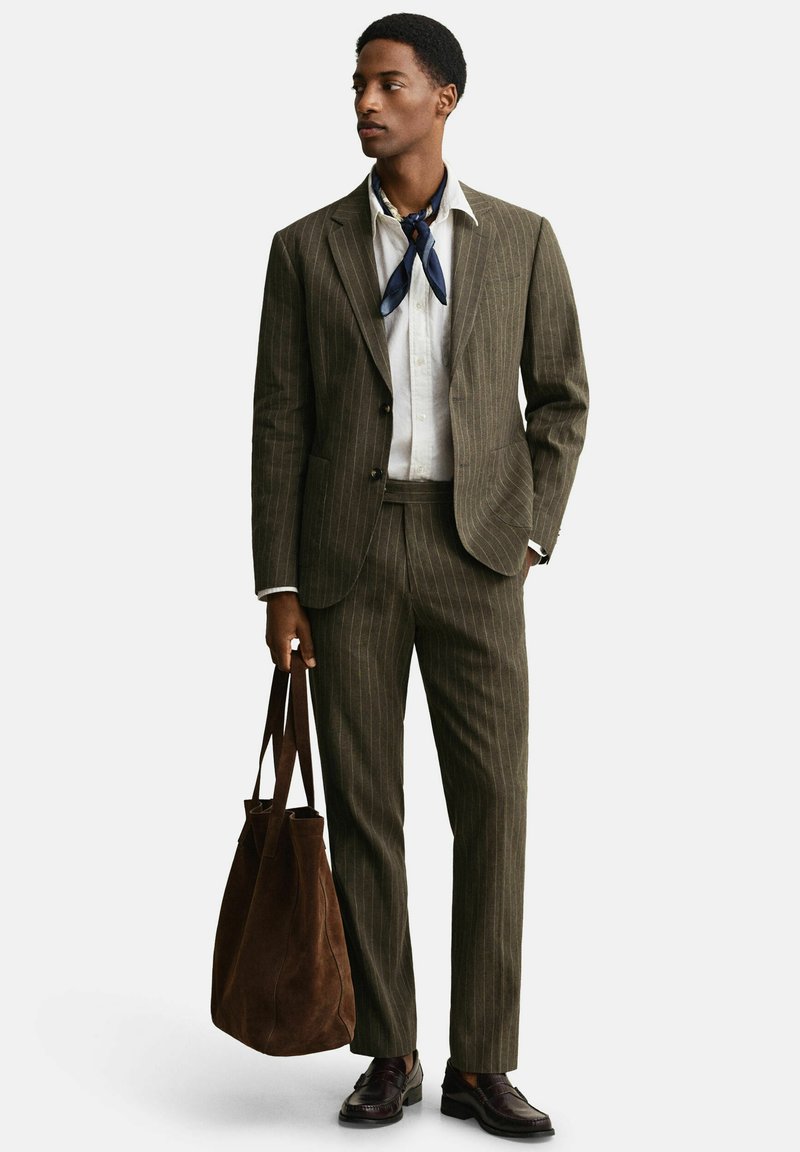 Man draagt een bruin krijtstreeppak en wit overhemd met marineblauwe sjaal, houdt een grote bruine suède tas vast, staat en kijkt naar links.