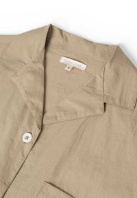 Hellbeige Hemd mit Kragen aus glattem Stoff, ausgestattet mit einer Brusttasche, einem weißen Knopf und einem Label mit "STUDIO FEDER" sowie der Größe "S".