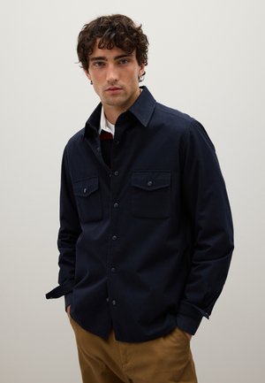 Young man with curly hair wears a dark navy button-up shirt with chest pockets over a white undershirt, standing against a plain light background.