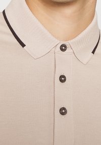 T-shirt polo beige avec un tissu texturé, trois boutons foncés et un contour noir contrastant au niveau du col. Vue rapprochée.