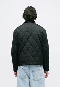 Chaqueta negra acolchada con un patrón de diamantes, que presenta un cuello acanalado y un corte ajustado. La tela parece suave y ligera.