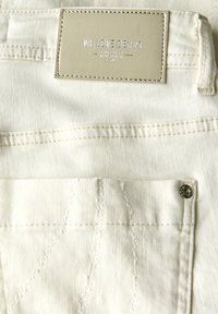 Vita denimjeans med en rektangulär etikett som står "VI ÄLSKAR DENIM" sydd på midjan. Har synlig sömnad och en metallknapp.