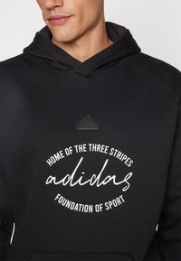 Felpa nera con cappuccio e tasca frontale, caratterizzata da un logo testurizzato e testo bianco: "HOME OF THE THREE STRIPES" e "FOUNDATION OF SPORT."