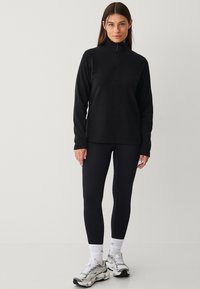 Zwarte fleece pullover met een kwart rits, lange mouwen en een ontspannen pasvorm, gecombineerd met zwarte leggings en sportieve schoenen.