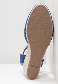 Sandale bleu en forme de coin avec tige en tissu, plateforme tissée de style espadrille, semelle en caoutchouc et clous décoratifs en ton argent.