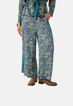 Conjunto floral de dos piezas con base verde azulado, mangas largas con un lazo en la cintura, y pantalones de pierna ancha con aberturas, combinados con botas marrones.