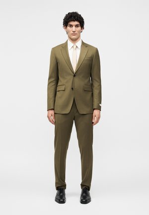 Άνδρας με σγουρά μαλλιά που φοράει μια πράσινη ελιά κοστούμι, λευκό πουκάμισο, μπεζ γραβάτα και μαύρα επίσημα παπούτσια, στέκεται μπροστά σε απλό ανοιχτό φόντο.