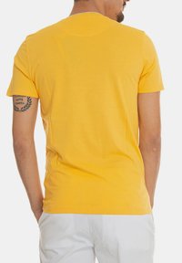 T-shirt in cotone giallo con maniche corte, collo rotondo e una texture liscia. Presenta un tatuaggio visibile sul braccio sinistro.