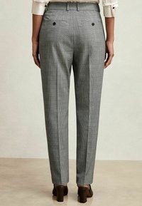 Mujer vestida con pantalones de cuadros grises y corte a medida, con bolsillos traseros abotonados, blusa blanca y zapatos de tacón cuadrado marrones, de pie sobre un suelo claro.