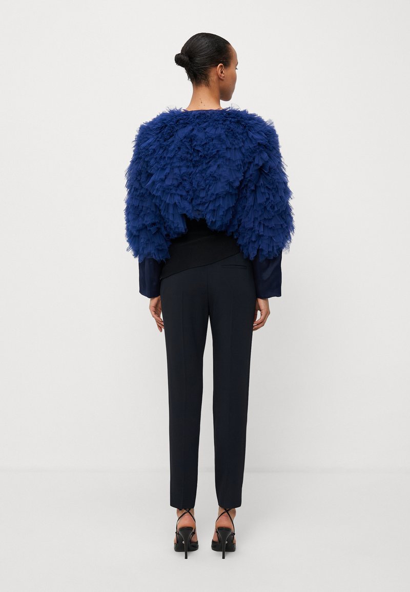 Granatowy teksturowany peleryna z warstwowym frędzlami na dopasowanej czarnej bluzce i dopasowanych czarnych spodniach, zestawiona z czarnymi sandałkami na obcasie.