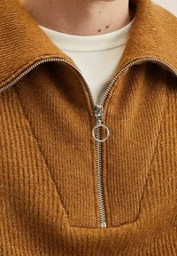 Brauner Strickpullover mit Reißverschluss, mit strukturierter Strickoptik, V-Ausschnitt und silbernem Reißverschluss. Der Kragen ist breit und formstabil.