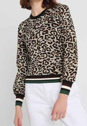 Femme portant un pull à manches longues à imprimé léopard avec des poignets et un ourlet rayés noir, blanc et vert, associé à un pantalon blanc.