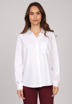 Chemise blanche à col, à manches longues, avec une poche sur la poitrine gauche et une texture lisse, portée avec un pantalon bordeaux.