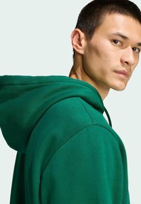 Zelená mikina s kapucňou na šnúrku, mäkká textúra a šité detaily, zobrazená z boku s detailným pohľadom na golier a rameno.