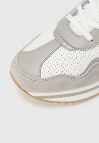 Close-up af grå og hvide perforerede sneakerspids med ruskind og læderpaneler samt hvide snørebånd på hvid baggrund.