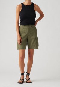 Pantalones cortos de carga verde oliva con bolsillos laterales y cierre de botón, combinados con una camiseta sin mangas negra y sandalias negras de tiras.