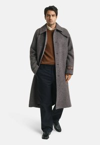 Lange, geruite wollen jas in bruin- en grijstinten, met een brede kraag en een opgestikte zak. Draagt over een bruine trui en donkere broek.