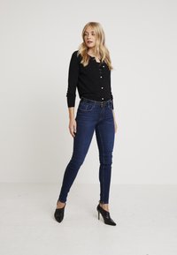 Černý dlouhý kardigan s kulatým výstřihem a knoflíkovými detaily, kombinovaný s tmavě modrými skinny džínami a černými vysokými podpatky.