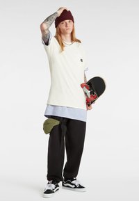 Camiseta corta de manga corta color crema con un bolsillo, superpuesta a una camisa más larga de color azul claro; pantalones de chándal negros; riñonera verde; patineta negra; gorro oscuro.
