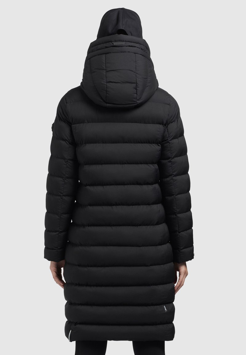 Eine Person trägt einen langen schwarzen, gesteppten Puffer-Mantel mit Kapuze und einer schwarzen Mütze, von hinten gegen einen schlichten Hintergrund gezeigt.