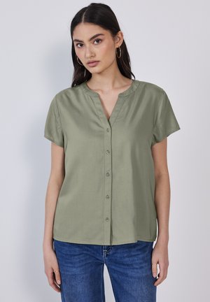 Vrouw met lang donker haar, die gouden oorringen, een kort-lang mouwen olijfgroen blouse met knopen en blauwe jeans draagt, staand tegen een effen achtergrond.