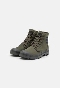 Bottes courtes en toile vert olive avec un capuchon en caoutchouc, semelle durable et lacets prononcés. Présente un patch logo circulaire sur le côté.