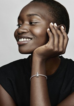 Silbernes Armband mit einem Anhänger, der eine kleine Figur zeigt, getragen an einem dunkelhäutigen Handgelenk. Der Hintergrund ist neutral und hebt den Schmuck hervor.