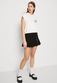 Valkoinen lyhyt hiha sweatshirt, jossa on logo, yhdistetty mustaan laskostettuun hameeseen, jossa on sivuaukot ja metalliset yksityiskohdat. Mustat ja valkoiset loafersit.