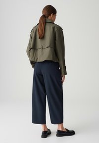 Olijf groene cropped jack met een rugklep en knoopdetail, gedragen met een wijde marineblauwe broek en zwarte instappers. Gladde stoftextuur.