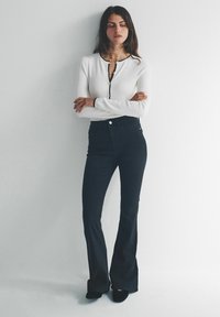 Bluză albă cu mâneci lungi și guler negru, asortată cu blugi înalți, evazați, de culoare închisă. Modelul stă într-un decor neutru.