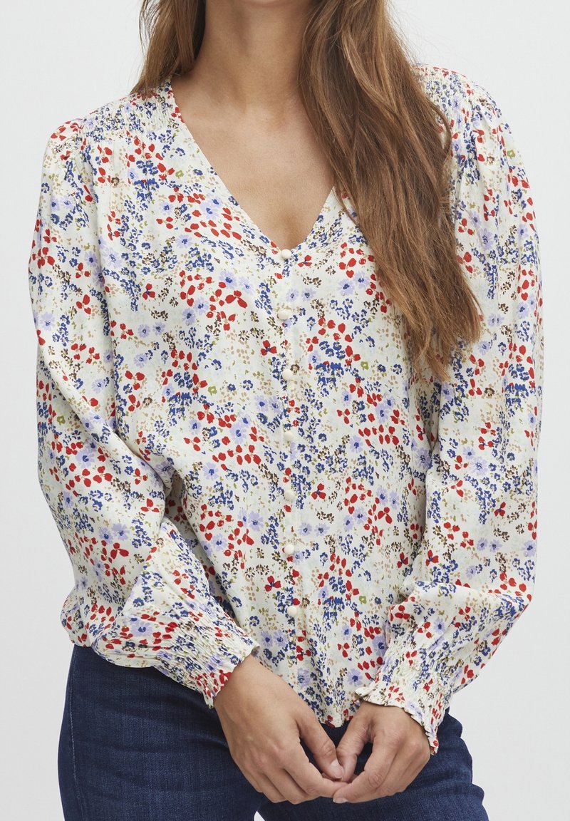 Bluse mit Blumenmuster in Creme, mit roten und blauen Blumen. V-Ausschnitt, Knopfverschluss und elastischen Bündchen. Leichter, weicher Stoff.