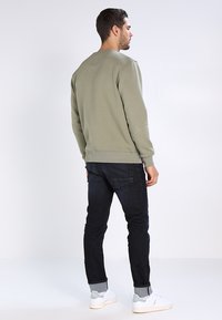 Sudadera verde oliva con cuello redondo y puños de canalé. Combinada con jeans oscuros y zapatillas blancas, mostrando una silueta casual.