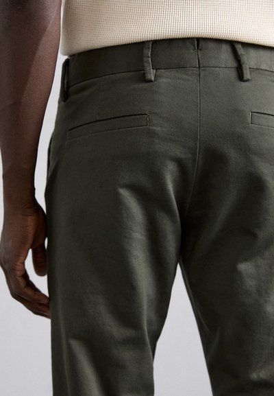 Grüne Baumwollhose mit glatter Textur, ausgestattet mit zwei Gesäßtaschen und Gürtelschlaufen, aus einem hinteren Winkel gezeigt.