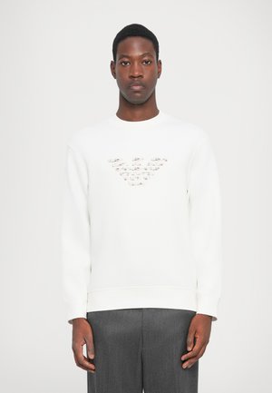White sweatshirt with a crew neckline and long sleeves. Features a textured logo design on the chest. Worn with gray tailored trousers.