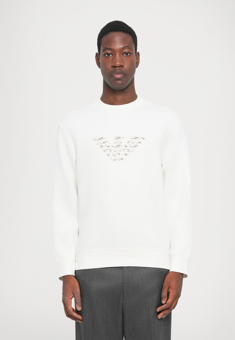 Sudadera blanca con cuello redondo y mangas largas. Presenta un diseño de logo texturizado en el pecho. Se lleva con pantalones sastre grises.