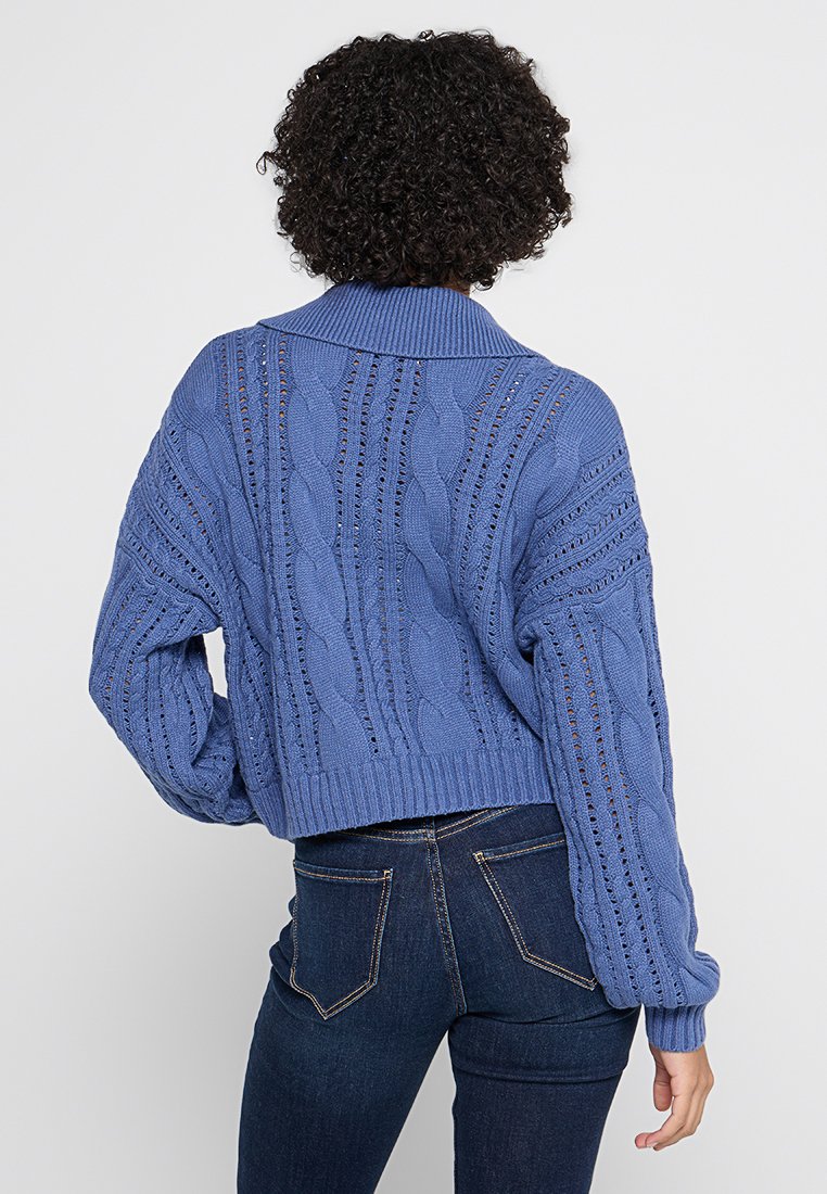 Maglione corto a maglia blu con colletto ampio, caratterizzato da motivi testurizzati e dettagli a tessitura aperta, abbinato a jeans scuri.