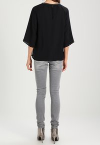 Schwarze, fließende Bluse mit Glockenärmeln und einem Schlüsselloch-Detail im Rücken. Kombiniert mit engen, hellgrauen Jeans und metallischen High Heels.