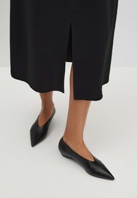 Schwarze Leder-Ballerinas mit spitzem Zehenbereich und niedrigem Absatz, kombiniert mit einem schwarzen Kleid mit einem Schlitz vorne.