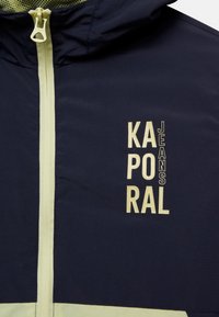 Granatowa kurtka przeciwwiatrowa z jasnozielonymi akcentami, wyposażona w centralny zamek błyskawiczny oraz pionowy napis "KAPORAL JEANS" w kontrastującym kremowym kolorze.