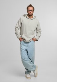 Sudadera gris clara con un bolsillo frontal y capucha con cordón, combinada con unos jeans holgados azul claro y zapatillas blancas.