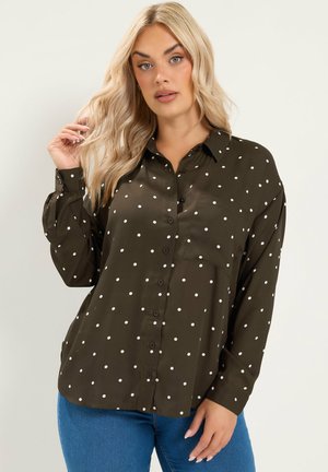 Vrouw met lang blond haar, gekleed in een olijfgroen overhemd met witte stippen en een blauwe spijkerbroek tegen een effen achtergrond.