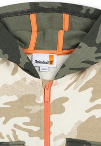 Camo-gepatroneerde hoodie met een tan en olijfgroene kleurenschema. Heeft een oranje rits, capuchon en een Timberland-label. Van een katoenen blend materiaal.